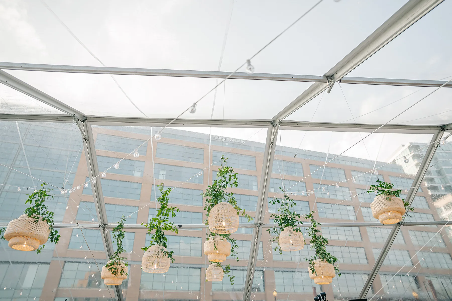 Hanging greenery and woven pendant lights inside a clear-top wedding tent