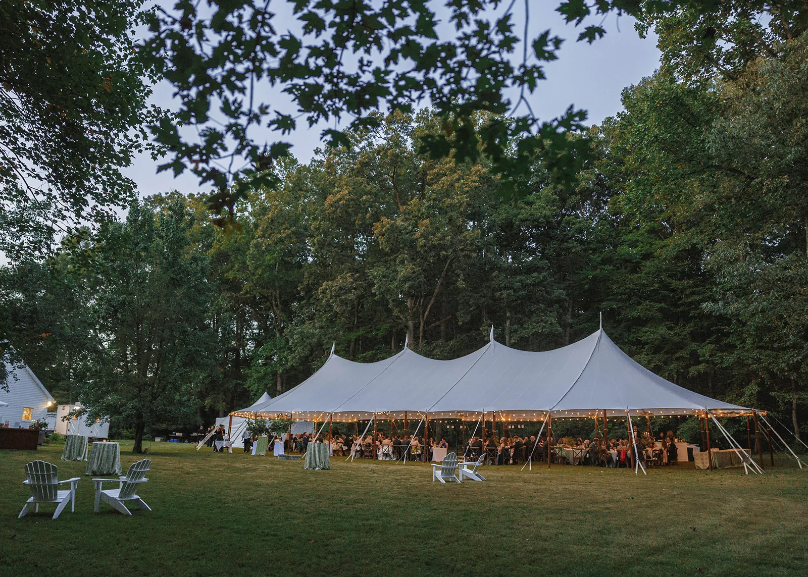 Lit sailcloth wedding tent on a wooded lawn at evening