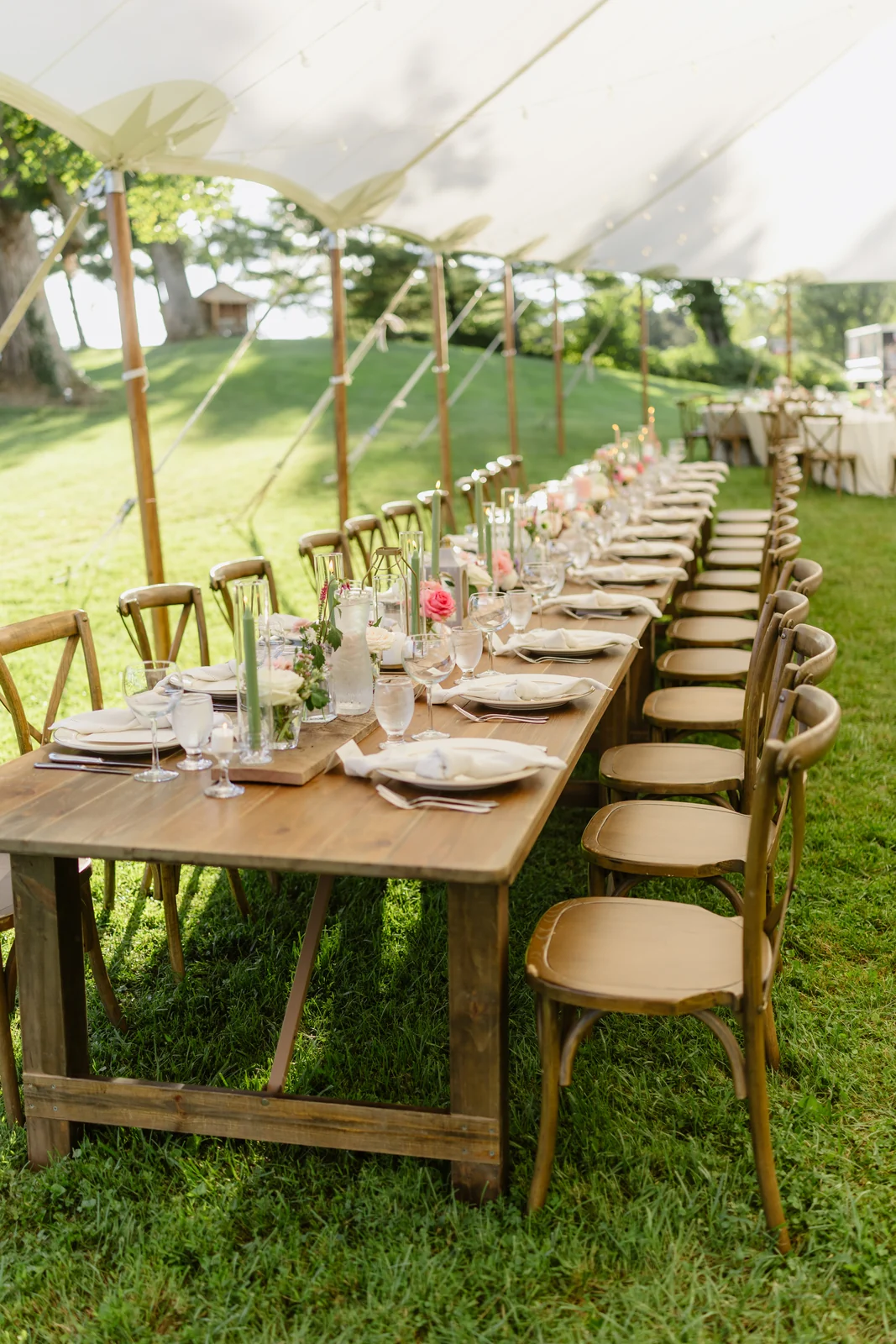 Long wooden reception table with floral arrangements and cross-back chairs under a sailcloth tent