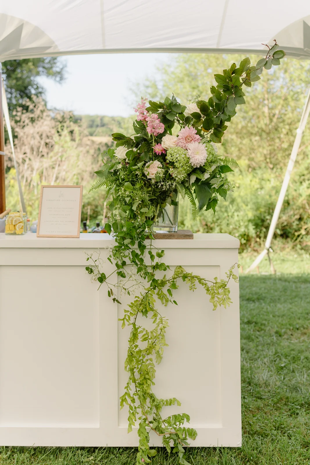 White wedding bar decorated with greenery and flowers beneath a tent